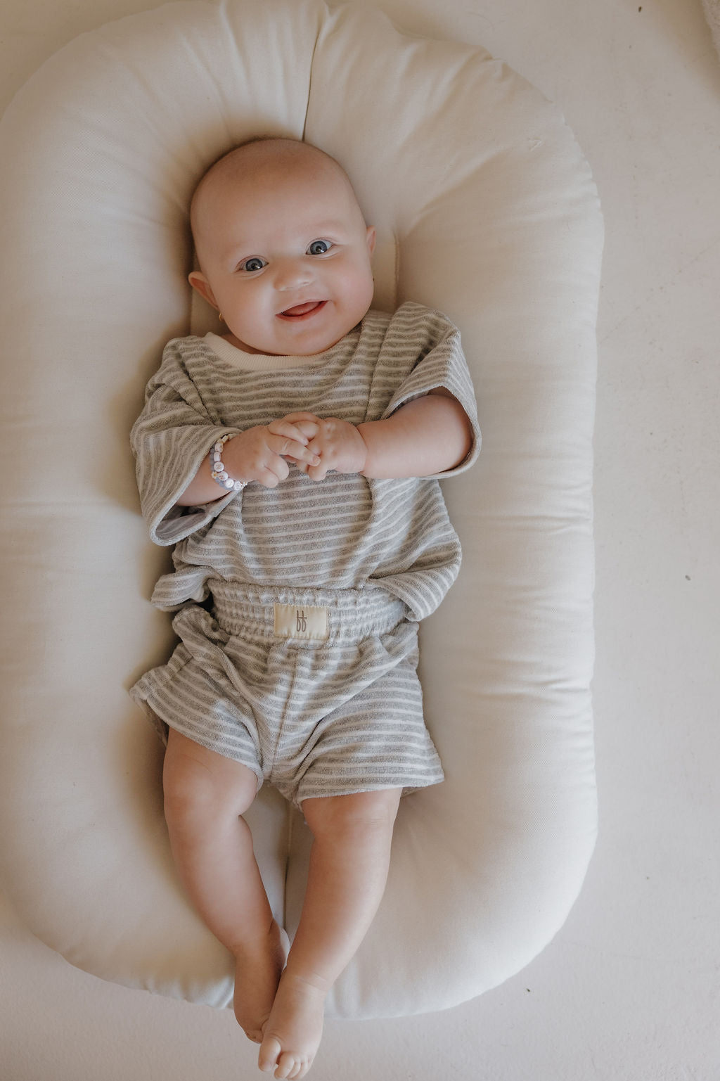 A smiling baby with blue eyes, wearing the forever french baby Child Terry Short Set in Grey Stripe, lies on a soft white cushion with hands clasped—capturing precious family moments while looking up at the camera.