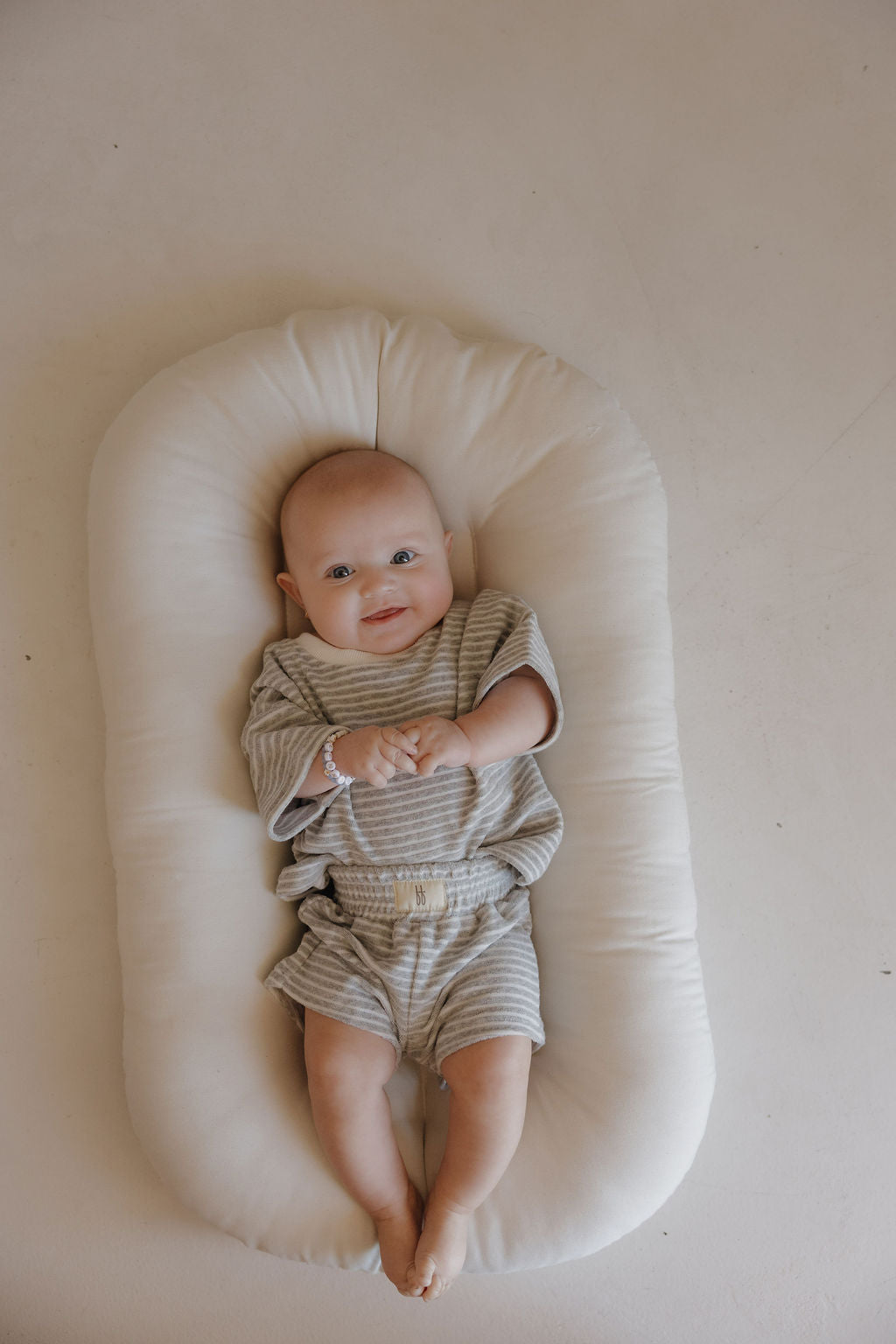 A baby in the forever french baby Child Terry Short Set | Grey Stripe lies on a soft, white oval cushion, smiling up and capturing a precious family moment against a plain light background.