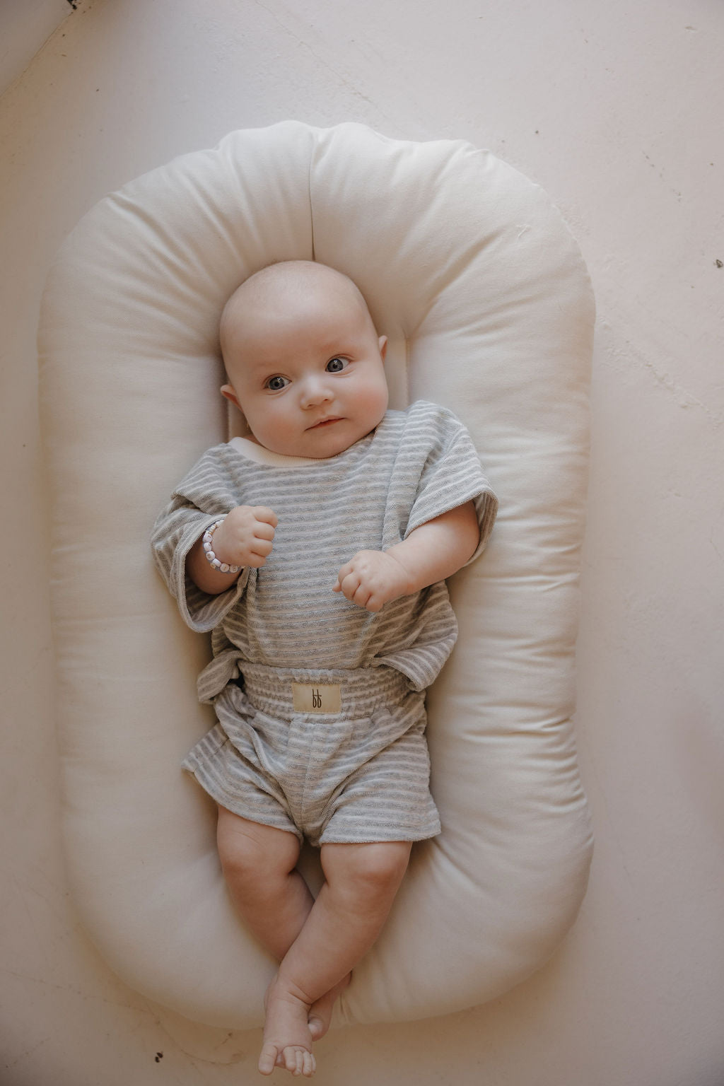 A baby in the forever french baby Child Terry Short Set | Grey Stripe lies on a soft, oval white cushion, gazing up sweetly—ideal for capturing precious family moments against a light backdrop.