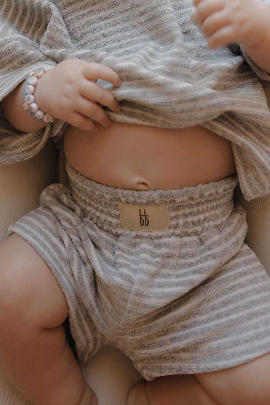 A baby in the forever french baby Child Terry Short Set | Grey Stripe lifts their shirt to reveal their belly and belly button, wearing a beaded bracelet—capturing sweet family moments.