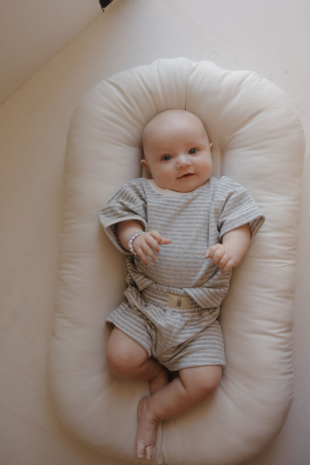 A baby wearing the forever french baby Child Terry Short Set in Grey Stripe lies on a soft cream cushion, smiling gently. Natural light highlights this sweet moment, perfect for capturing precious family memories.