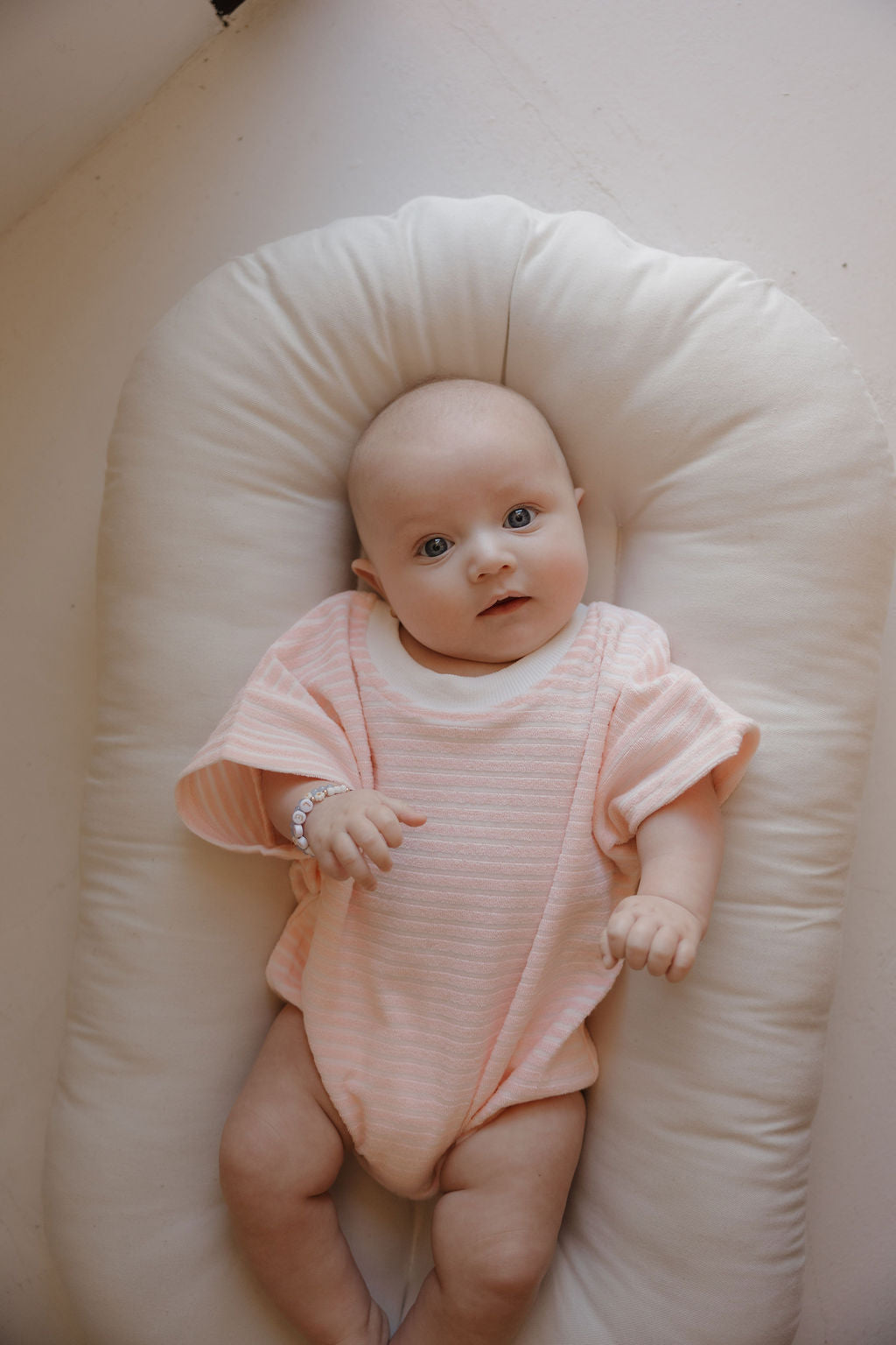 A light-skinned baby with blue eyes lies on a soft white cushion, wearing the forever french baby Terry T-Shirt Romper | Cotton Candy, looking up calmly—perfect for capturing sweet family memories.