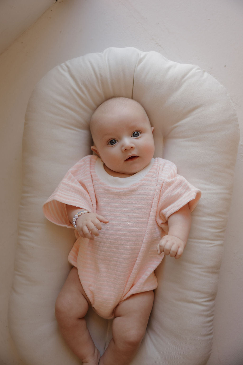 A baby wearing the forever french baby Terry T-Shirt Romper | Cotton Candy lies on a soft, white cushion, gazing up with wide eyes and hands relaxed—a sweet family moment. The romper's light pink hue adds to the charm, and a bracelet adorns one wrist.
