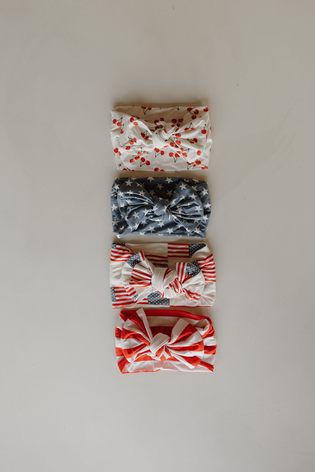 Four forever french baby Bamboo Head Wraps—including Sweetheart Cherries, blue stars, American flags, and red stripes—are arranged vertically on a neutral background. All wraps are crafted from soft bamboo for comfort.