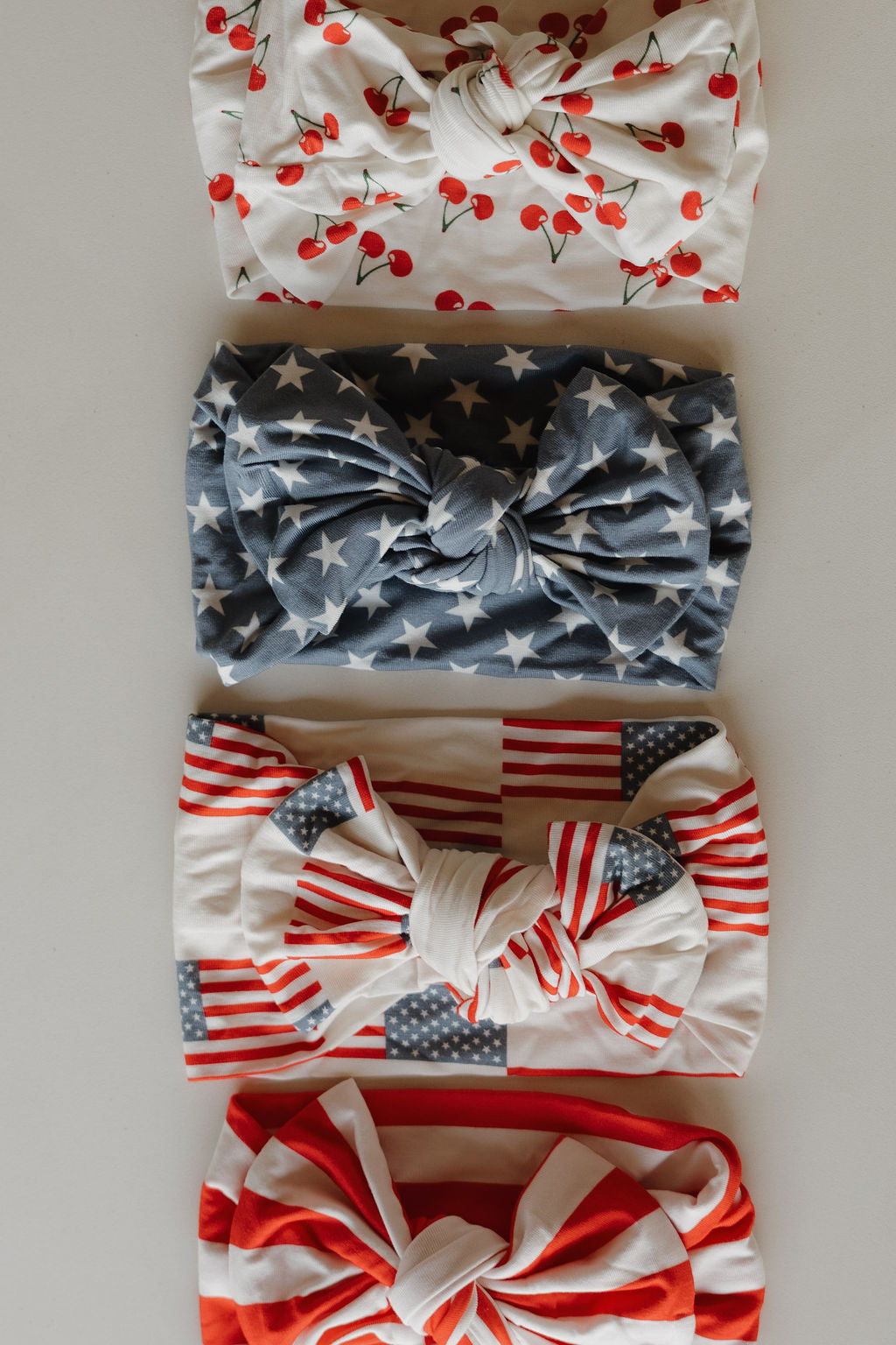 Four knotted Bamboo Headwraps from forever french baby are arranged vertically: white with red cherries, blue with white stars, red and white stripes with blue stars, and red and white stripes with red trim.