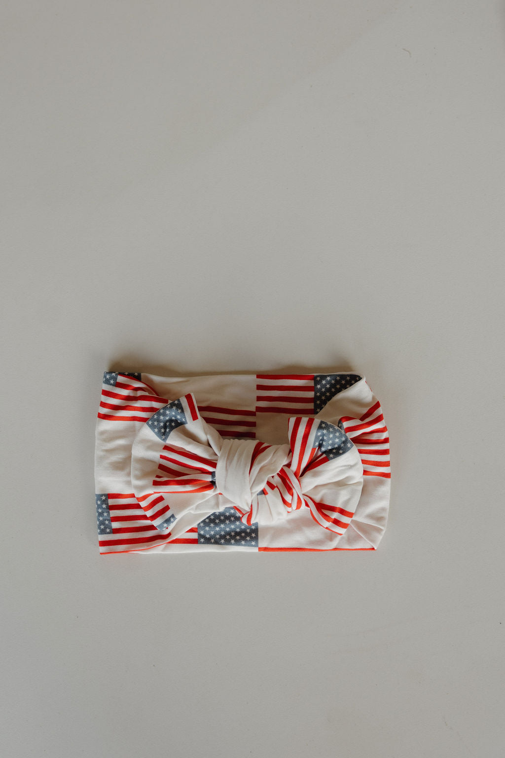 The forever french baby Bamboo Head Wrap | Americana, featuring red and white stripes with blue and white stars in an American flag-inspired pattern, is displayed laid flat on a plain light surface.