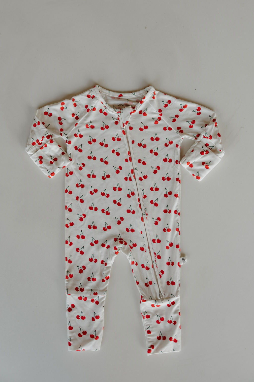 The Bamboo Zip Pajamas | Sweetheart Cherries by forever french baby is a white, long-sleeve onesie with a red cherry print and diagonal zipper, made from breathable fabric and shown laid flat on a light surface.
