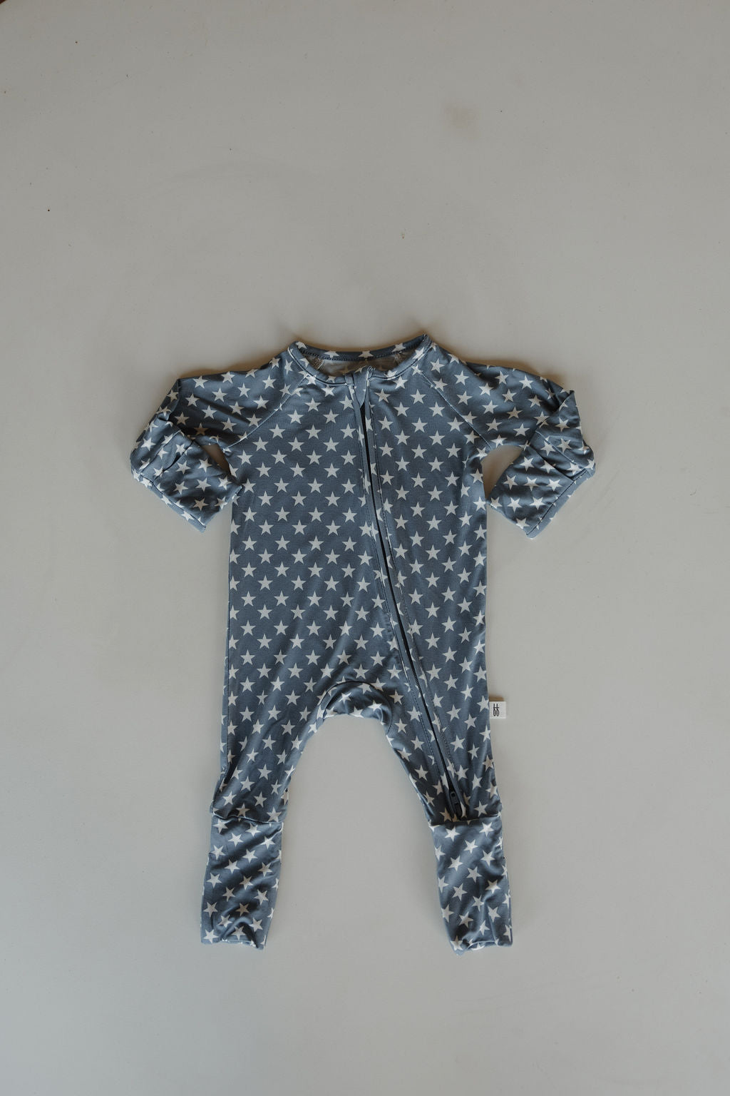 The forever french baby Bamboo Zip Pajamas | Star Spangled is a blue onesie with long sleeves and legs, featuring a white star pattern and a diagonal front zipper. Made from breathable fabric, it's shown flat on a light gray surface.