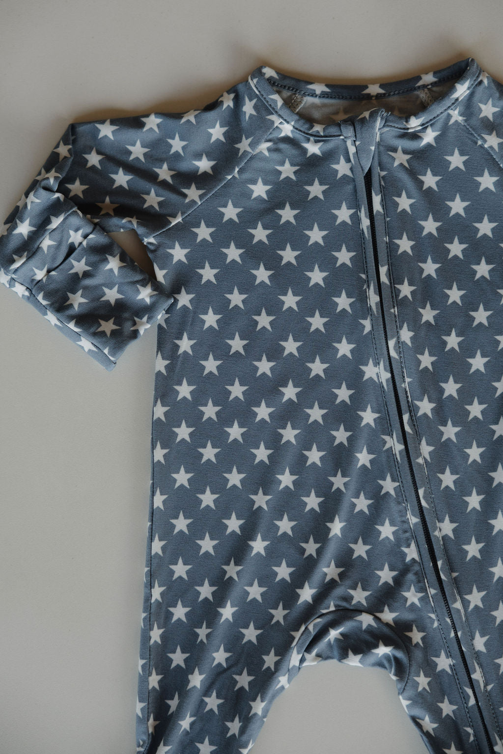 The forever french baby Bamboo Zip Pajamas | Star Spangled is a blue, long-sleeve baby onesie with a white star pattern and front zipper, made from breathable fabric and shown laid flat for a soft, comfy look.