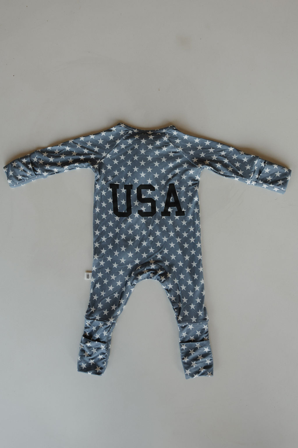 The forever french baby Bamboo Zip Pajamas | Star Spangled features a blue design with white stars and bold black "USA" text on the back, crafted from breathable fabric for all-day comfort, shown laid flat against a light surface.