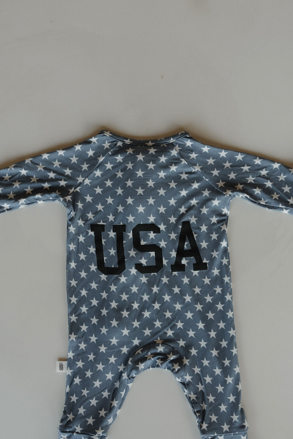 The forever french baby Bamboo Zip Pajamas | Star Spangled features a blue, long-sleeve design with white stars and bold black "USA" on the back, offering breathable comfort and laid flat for a cozy look.
