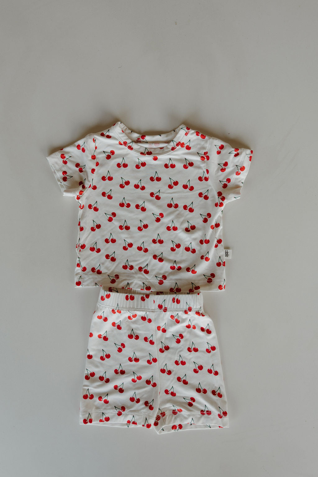 The forever french baby Bamboo Short Set | Sweetheart Cherries features a matching kid's short-sleeve shirt and shorts in white with a red cherry print, crafted from breathable fabric and shown flat on a light background.