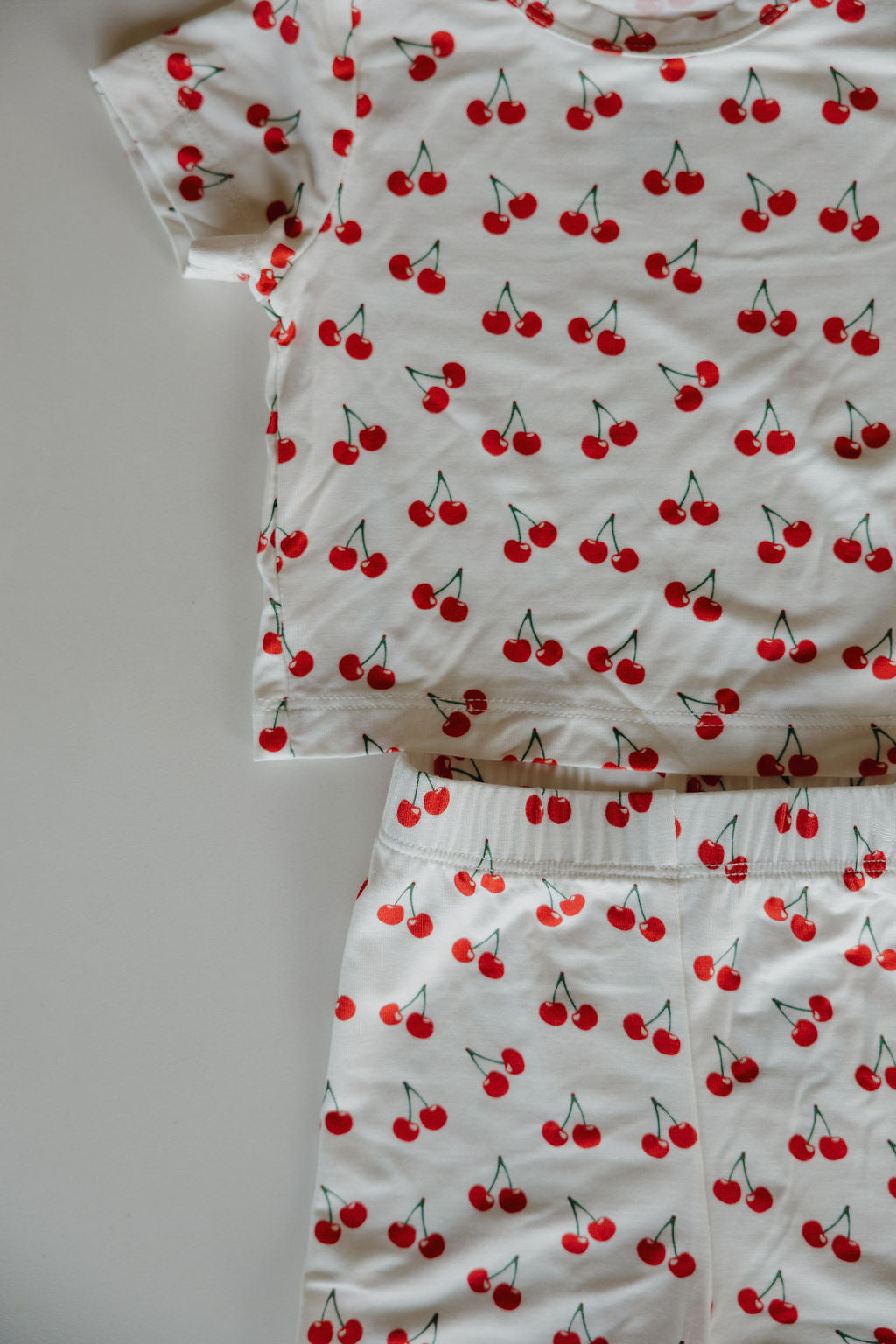 The Bamboo Short Set | Sweetheart Cherries by forever french baby is a white, short-sleeve pajama set with a red cherry print, made from breathable fabric, including a top and matching bottoms shown on a light surface.