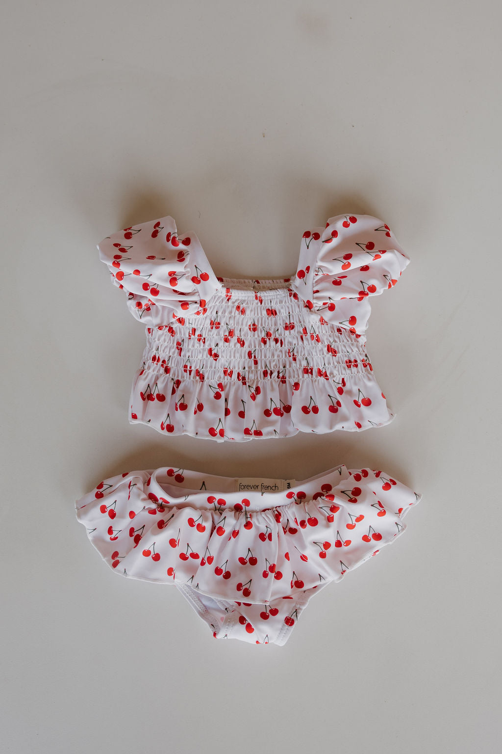 The forever french baby Toddler Two Piece Swimsuit | Sweetheart Cherries features a white smocked top with puffed sleeves and matching cherry-print bottoms, both displayed on a light surface.