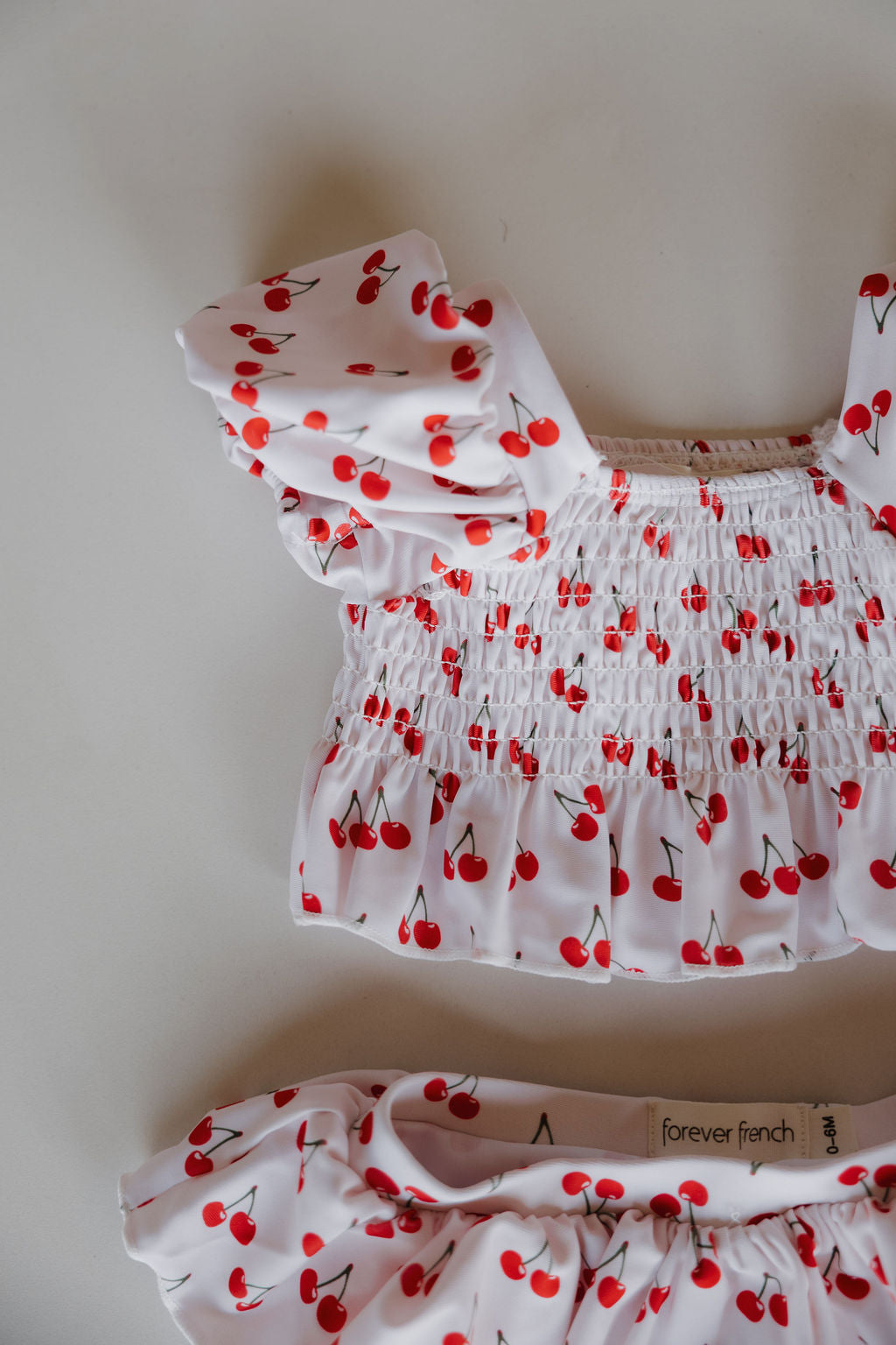 A white, short-sleeved Toddler Two Piece Swimsuit | Sweetheart Cherries by forever french baby features a playful red cherry print and is shown on a flat surface with the brand label visible inside the waistband.