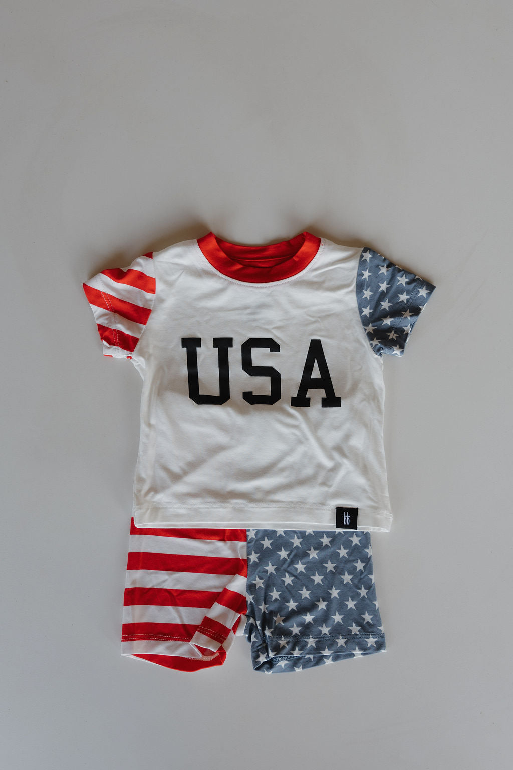 The forever french baby Bamboo Short Set | Stars & Stripes is a kids' pajama set with a white "USA" top, red collar, striped and star-patterned sleeves, and matching shorts—one leg striped, one star-patterned—crafted from breathable bamboo fabric.