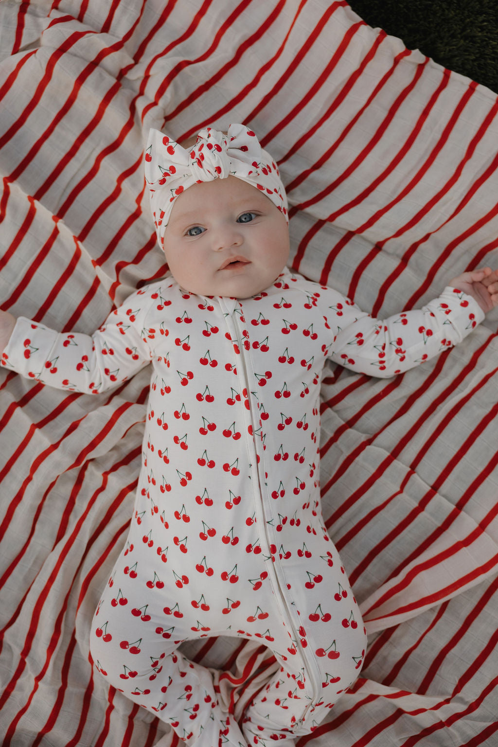 A baby in white wears a forever french baby Bamboo Head Wrap | Sweetheart Cherries, lying on a white blanket with red stripes and looking up with a neutral expression.