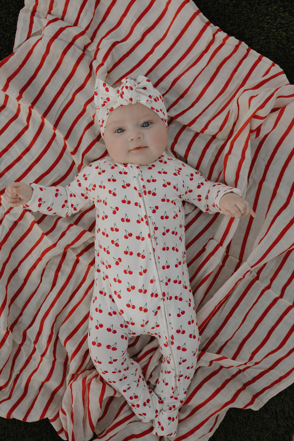 A baby in forever french baby Bamboo Zip Pajamas | Sweetheart Cherries, featuring cherry prints and a matching headband, lies comfortably on a red and white striped blanket thanks to the breathable fabric.