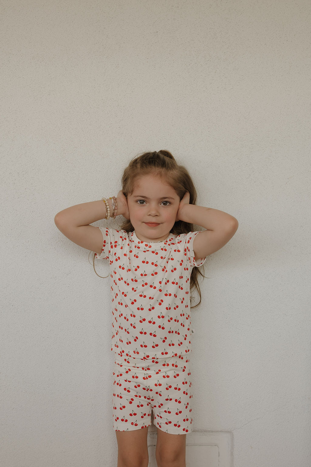 A young girl with light brown hair in a half-up style stands against a white wall, wearing the forever french baby Bamboo Short Set | Sweetheart Cherries, featuring a cherry print and made from breathable fabric.
