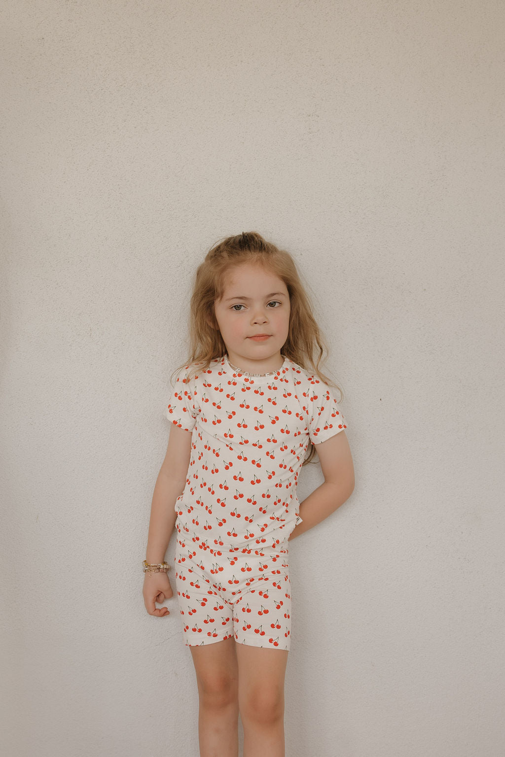 A young girl with long blonde hair stands against a plain light wall, wearing the forever french baby Bamboo Short Set | Sweetheart Cherries—breathable sleepwear featuring a white T-shirt and shorts with a red cherry print. Her left hand rests on her hip.