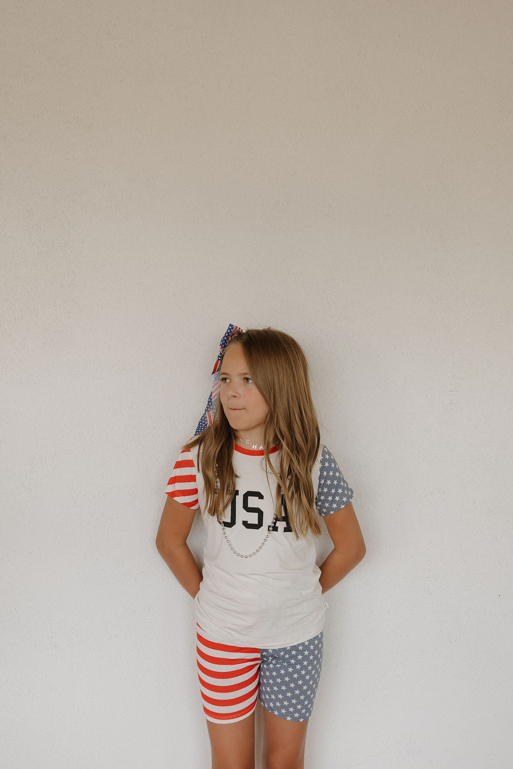 A young girl stands against a white wall, wearing the forever french baby Bamboo Short Set | Stars & Stripes, featuring breathable fabric with red and white stripes on one side and blue with white stars on the other, plus a beaded necklace and hair bow.
