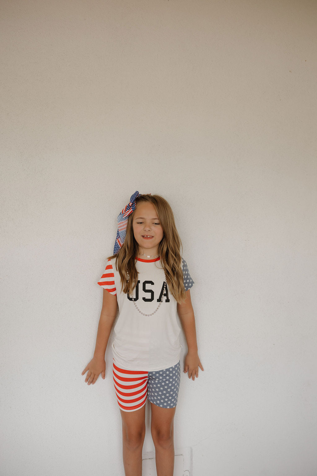A young girl smiles with eyes closed, wearing the forever french baby Bamboo Short Set | Stars & Stripes, featuring a USA-themed shirt and matching red striped and starry shorts, finished with a red, white, and blue bow.