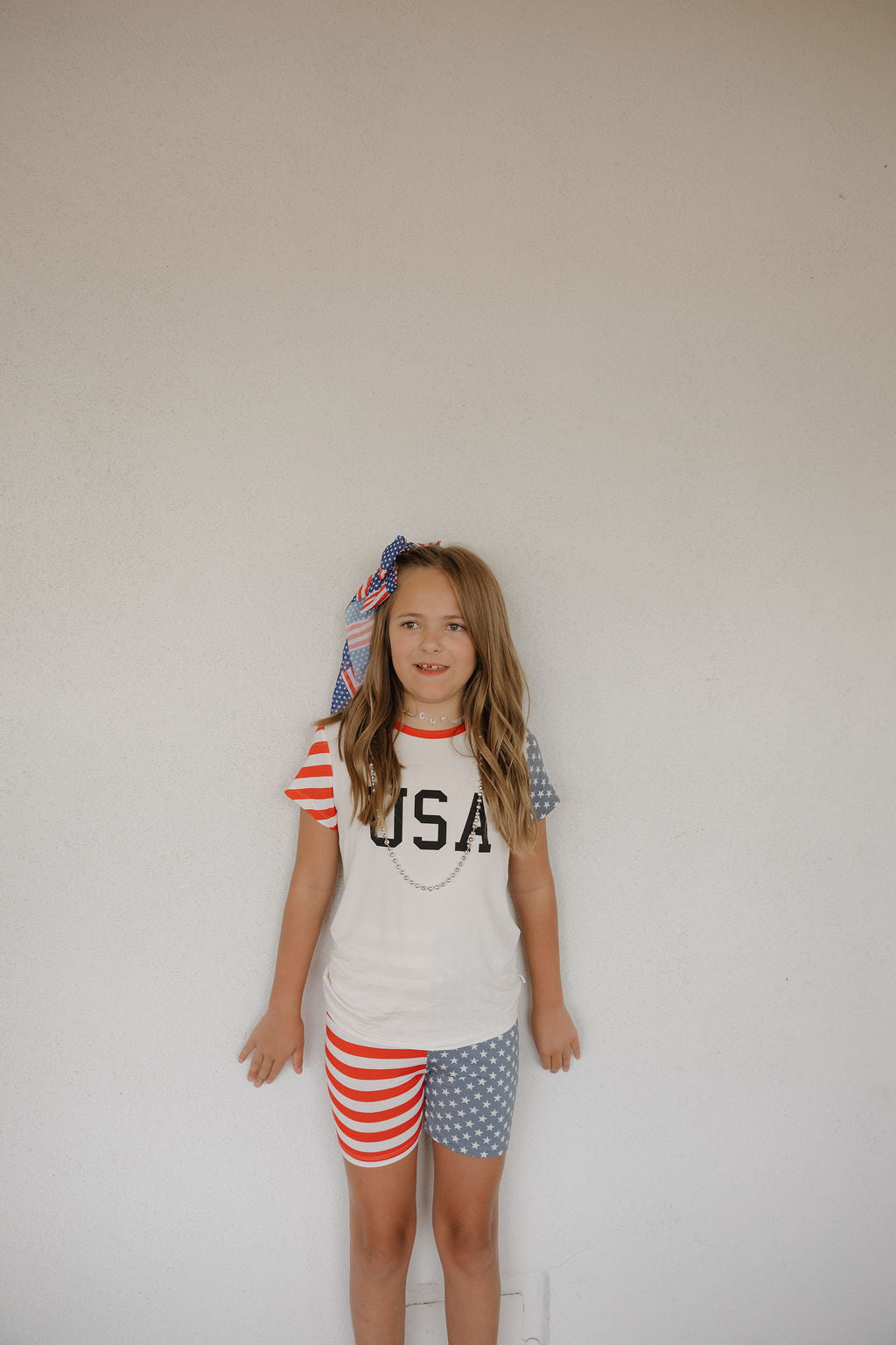 A young girl with long blonde hair and a festive bow stands against a white wall, wearing the forever french baby Bamboo Short Set | Stars & Stripes, featuring "USA" on the top and red-striped, blue star shorts in breathable fabric.
