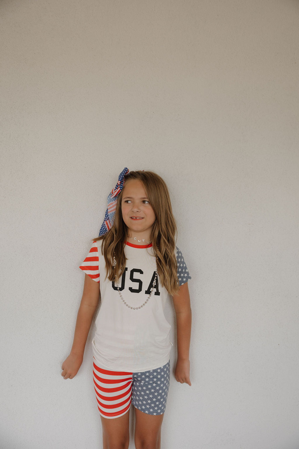 A young girl with long hair and a festive bow wears the forever french baby Bamboo Short Set | Stars & Stripes, featuring breathable fabric, “USA” on the shirt, red/white stripes on one sleeve and leg, and blue with white stars on the other.