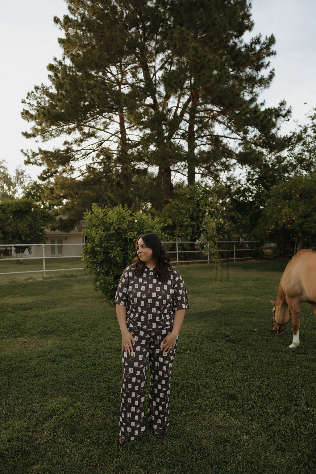 Wearing the forever french baby Women's Boxy Tee Lounge Set | With Love from AZ, a woman stands on grass in a fenced yard with trees as a light brown horse grazes nearby.