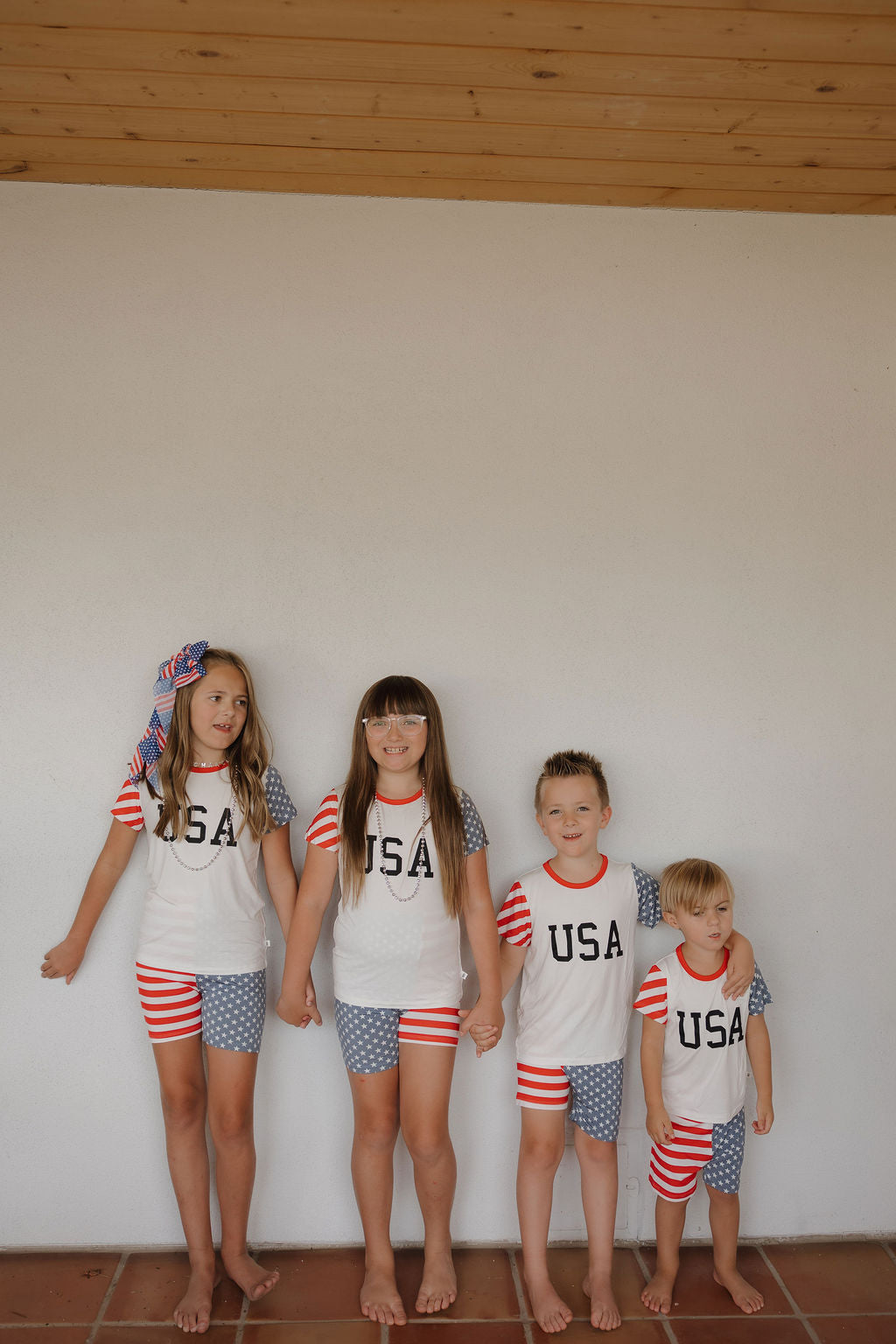 Four children smile in matching forever french baby Bamboo Short Set | Stars & Stripes outfits with "USA" on their shirts. Made from breathable fabric, their comfy attire includes a large striped bow in the oldest girl's hair.