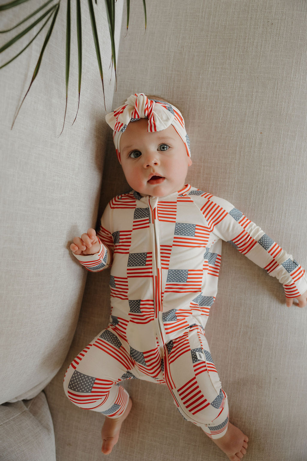 A baby in a forever french baby Bamboo Head Wrap | Americana and a stars-and-stripes onesie relaxes on a light-colored couch, gazing upward with one arm bent while a green leafy plant hangs into the scene.
