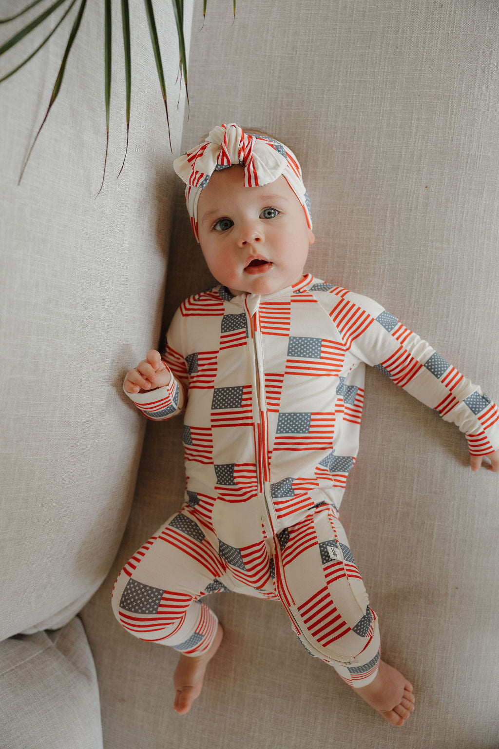 A baby in forever french baby's Bamboo Zip Pajamas | Americana, featuring American flag prints, lies on a light-colored couch wearing a headband and looking up curiously. A few leaves appear in the upper left corner.