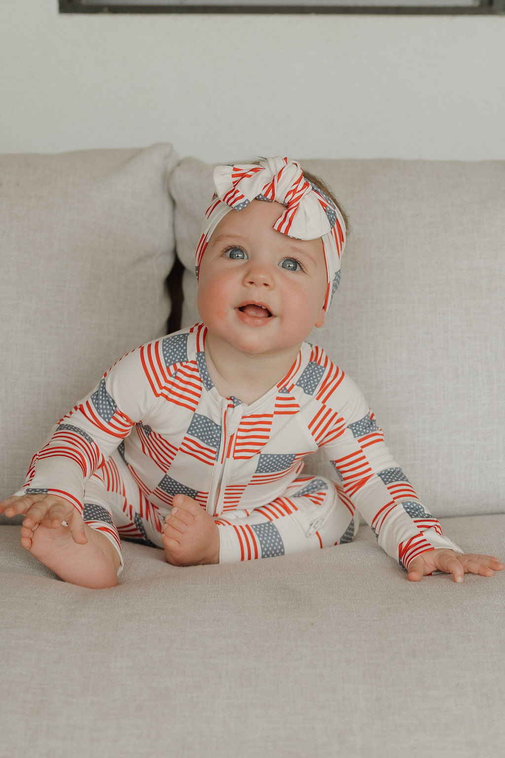 A baby wearing the forever french baby Bamboo Head Wrap | Americana sits on a light-colored couch, smiling and gazing upward. The stars and stripes head wrap completes the patriotic look.
