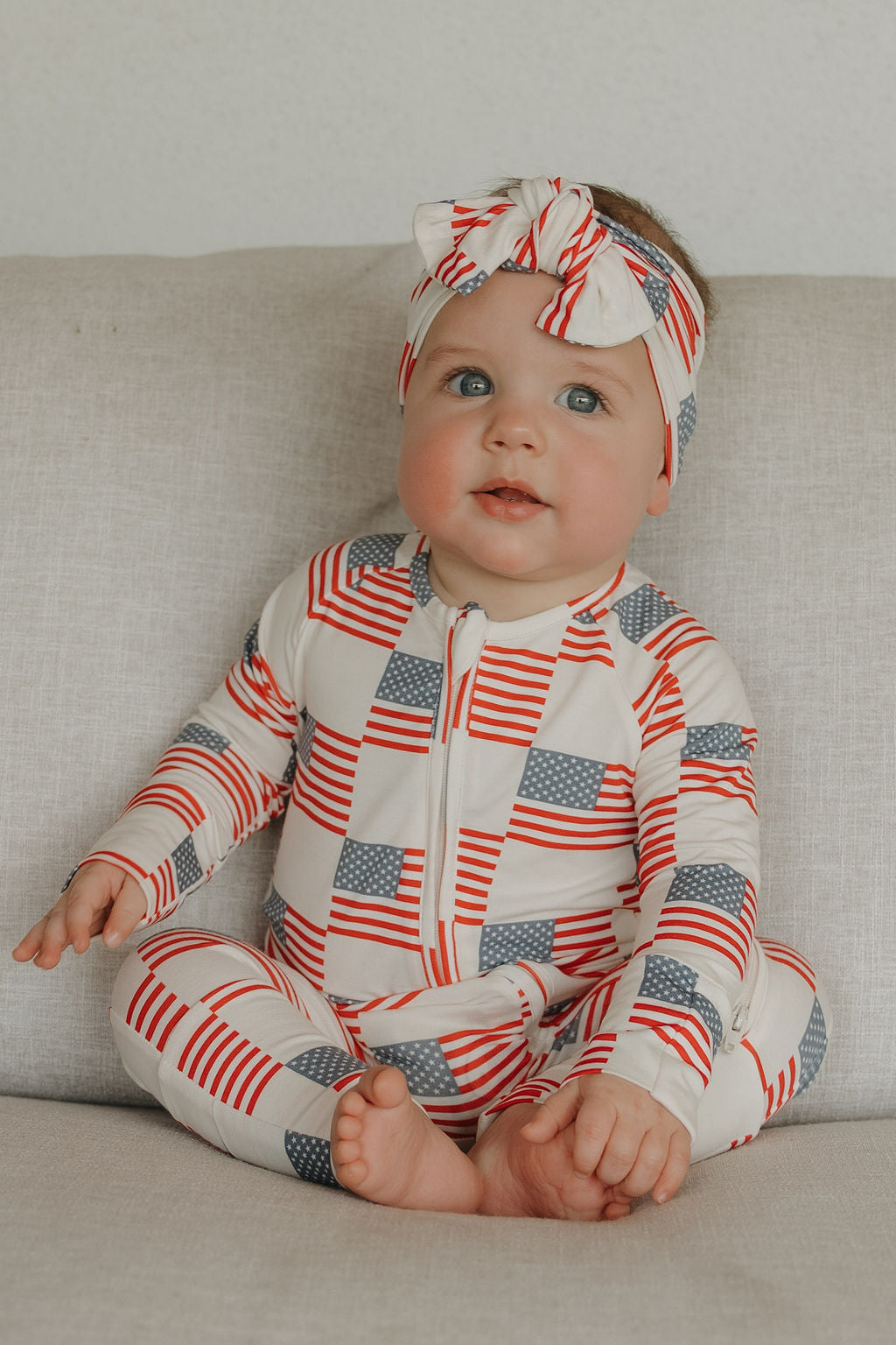 A baby in a forever french baby Bamboo Head Wrap | Americana and a red, white, and blue zip-up outfit sits on a light-colored couch, looking up and smiling.