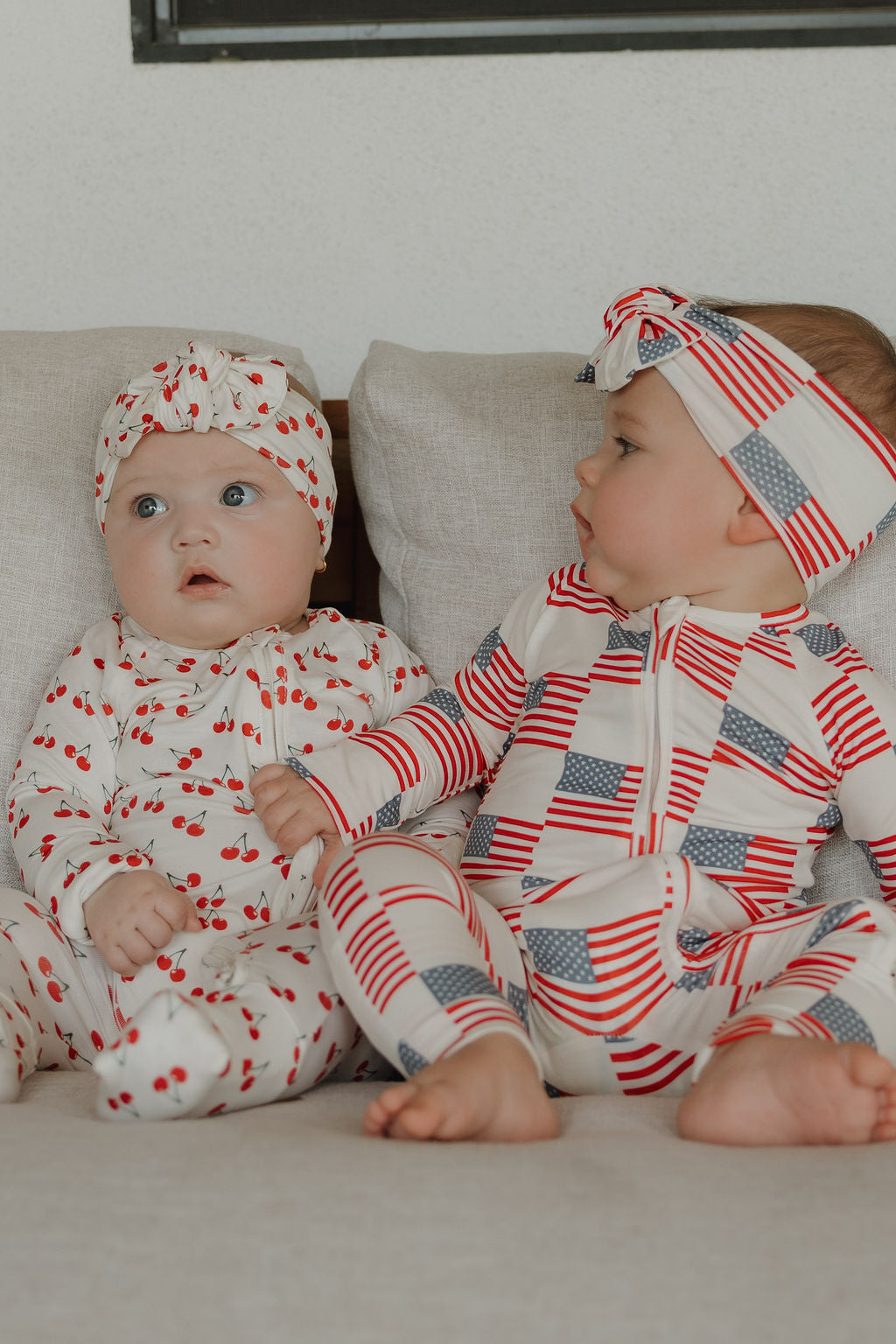 Two babies sit on a couch. One wears a white outfit with red cherries and a matching head wrap; the other wears a forever french baby Bamboo Head Wrap | Americana and a coordinating white outfit with American flags.