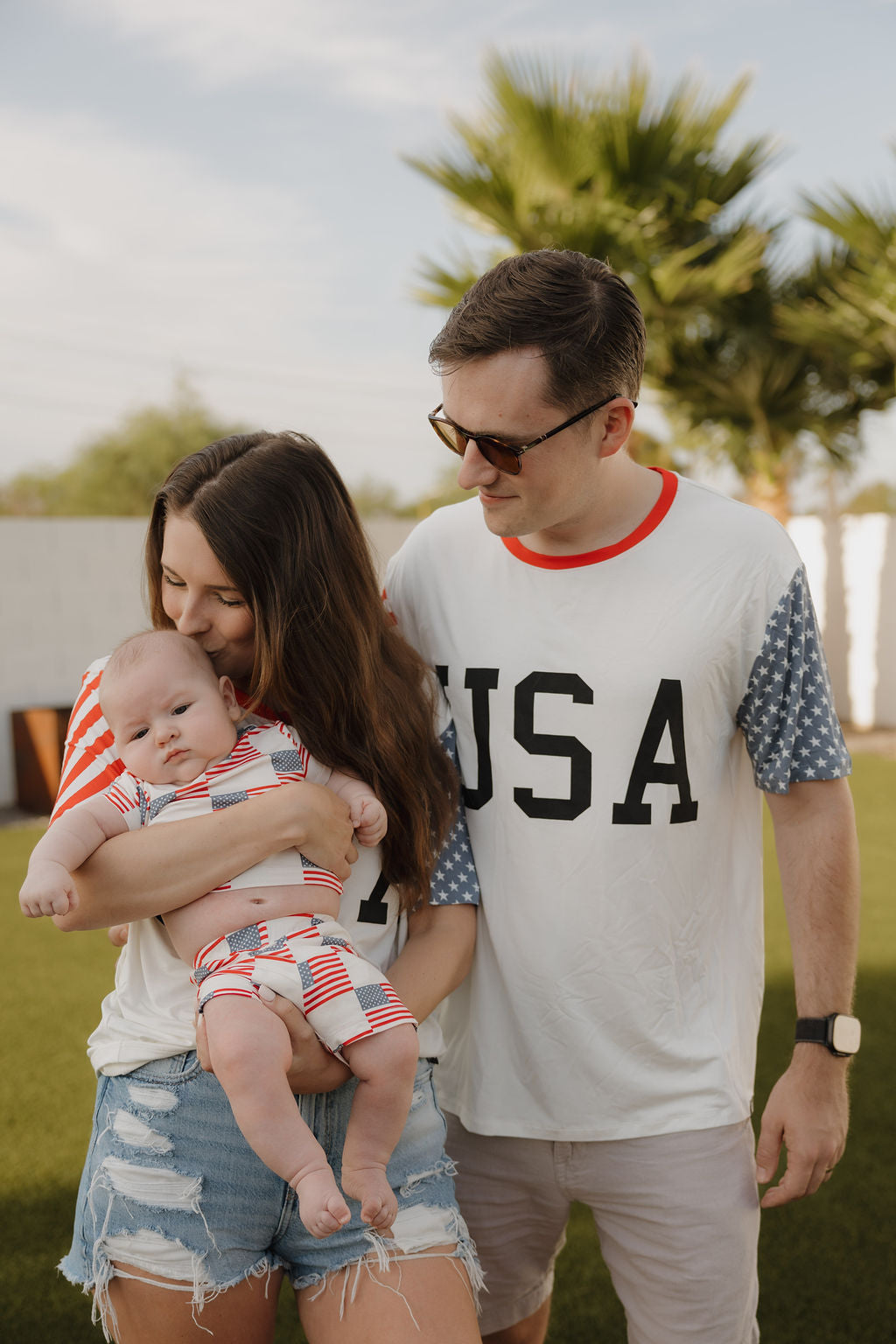 A woman holds a baby in patriotic bamboo clothing while a man beside her wears the forever french baby Adult Bamboo T-Shirt | Stars & Stripes. They stand outdoors on a sunny lawn with palm trees behind them.