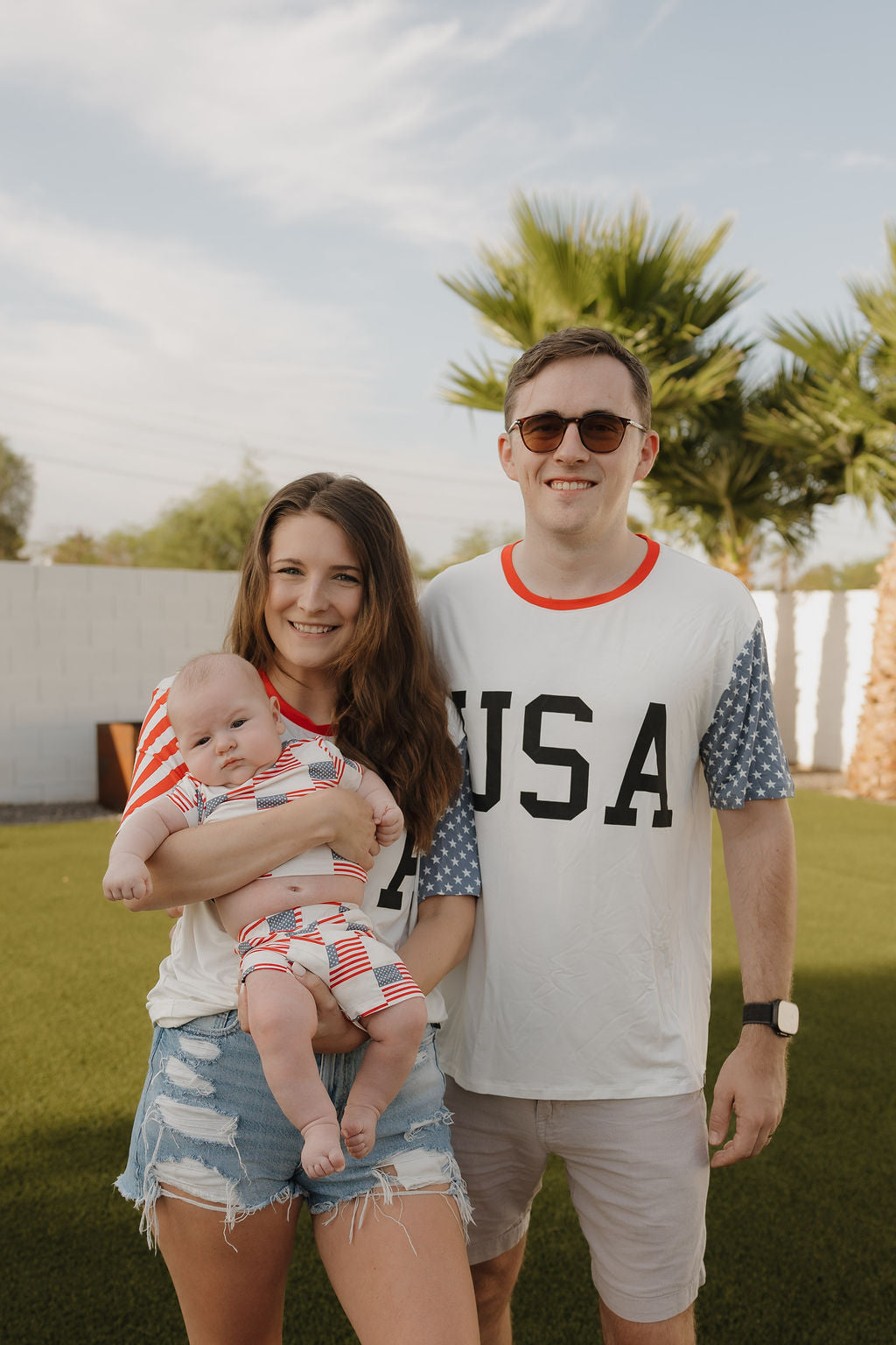 A woman holds a baby next to a man outdoors, all wearing forever french baby Adult Bamboo T-Shirt | Stars & Stripes with breathable fabric. Palm trees and a white wall are in the sunny background.