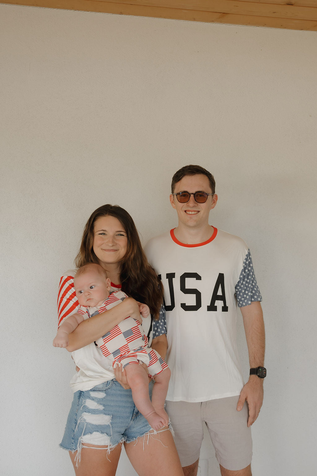 A smiling woman in breathable fabric holds a baby, both wearing red, white, and blue. Beside them is a man in the forever french baby Adult Bamboo T-Shirt | Stars & Stripes. All are posed before a plain light-colored wall.