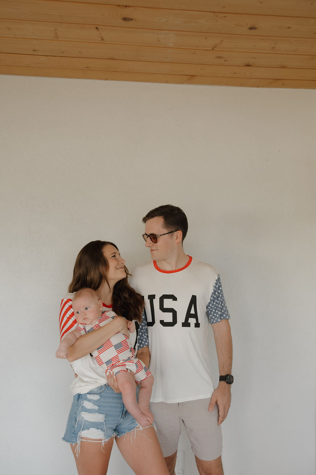 A woman holding a baby and a man stand together indoors, smiling. The man wears the forever french baby Adult Bamboo T-Shirt | Stars & Stripes, featuring breathable fabric and bold red, white, and blue patterns with "USA" on the front.