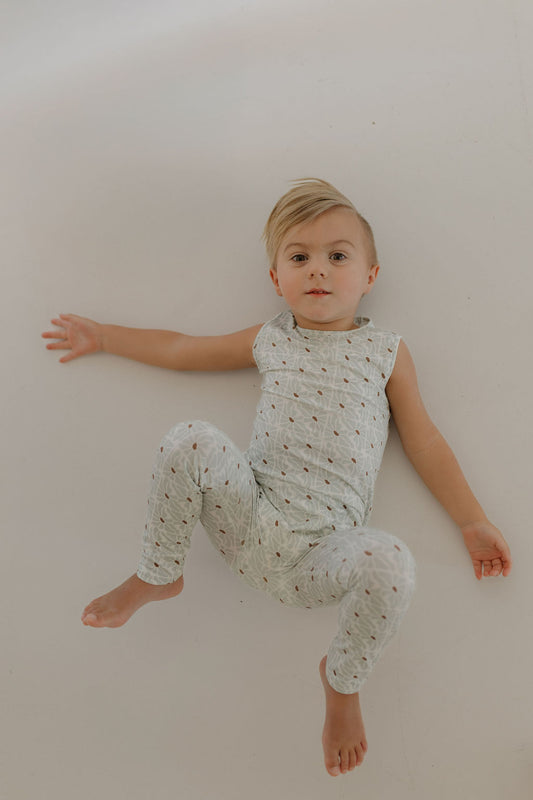 A small child with short, light hair lies on a white surface, dressed in a breathable, patterned Harem Bamboo Romper by forever french baby. One arm extends to the side as their bent legs rest comfortably. The child gazes into the camera with serene curiosity.