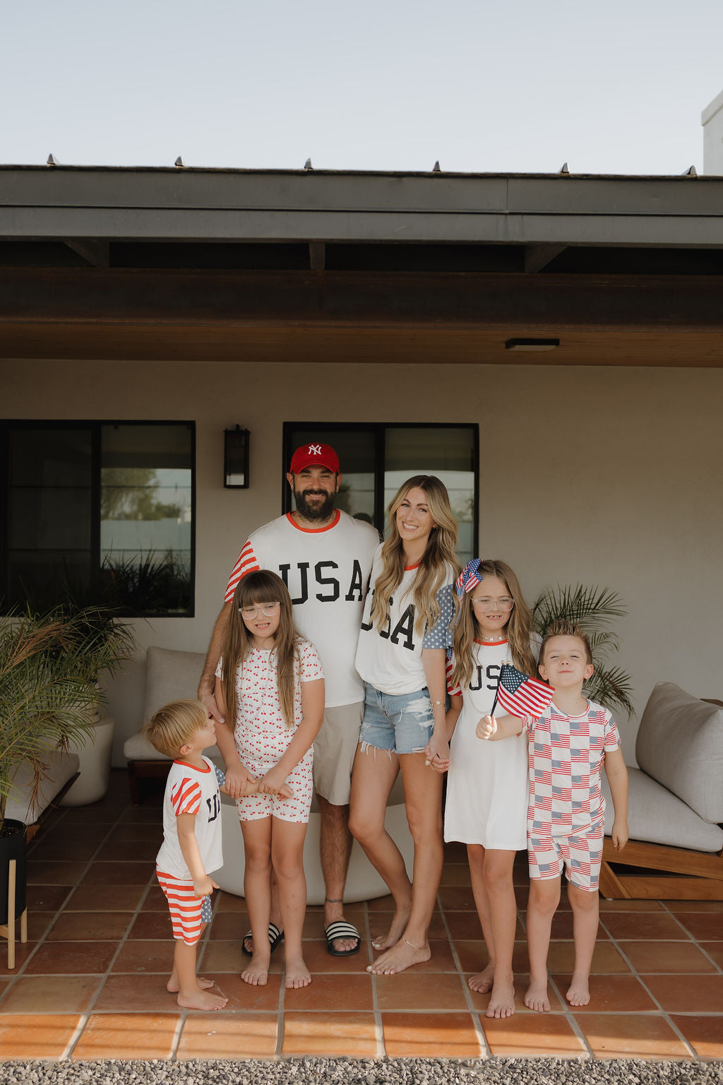 A barefoot family of six smiles on their patio, wearing forever french baby’s breathable, hypo-allergenic Bamboo T-Shirt Dress | Stars & Stripes in patriotic red, white, and blue.