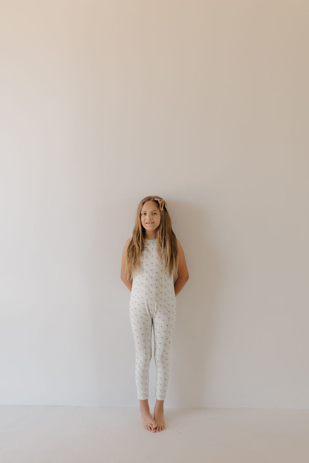 A young girl with long hair stands barefoot against a plain, light wall. She wears the Harem Bamboo Romper from forever french baby, featuring breathable fabric with a subtle pattern. Her hands are behind her back, and she looks calm.