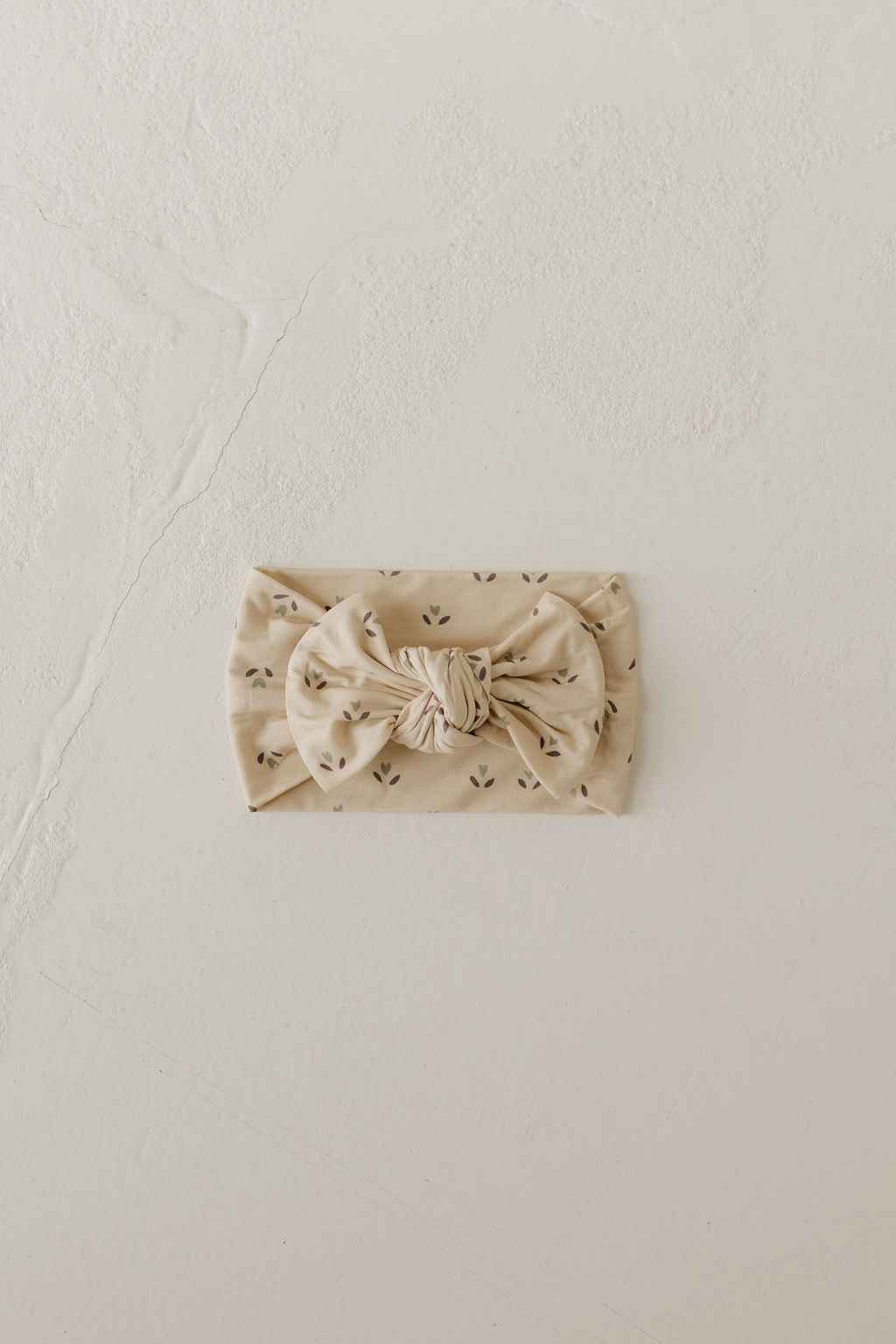 The Flora Bamboo Head Wrap from forever french baby, in beige with a central bow showcasing a subtle pattern of small black dots, lies elegantly on a light, textured surface.