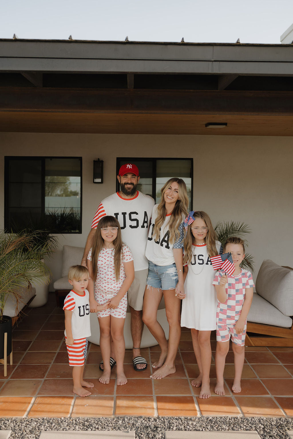 A family of five celebrates in breathable comfort, smiling on a porch in forever french baby’s Bamboo T-Shirt Dress | Stars & Stripes, holding American flags and dressed in patriotic red, white, and blue.
