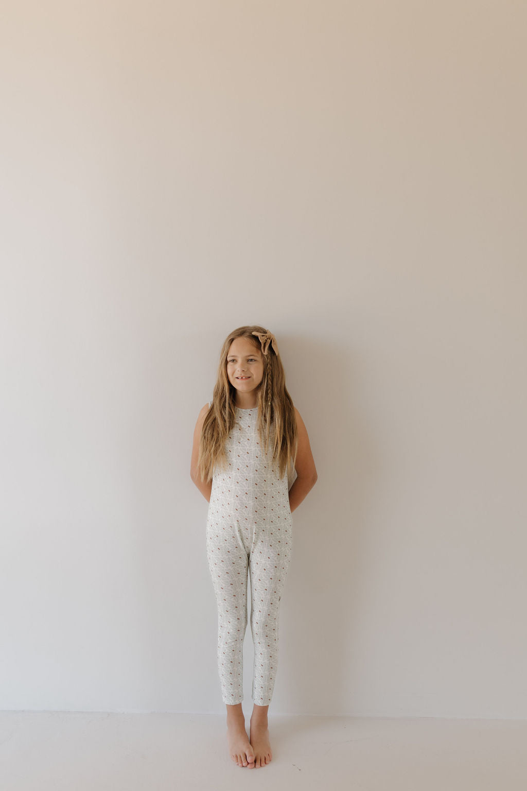 A young girl with long, light brown hair and a hairpin stands barefoot against a light-colored wall, wearing a sleeveless Harem Bamboo Romper from forever french baby. She keeps her hands behind her back and maintains a neutral expression.