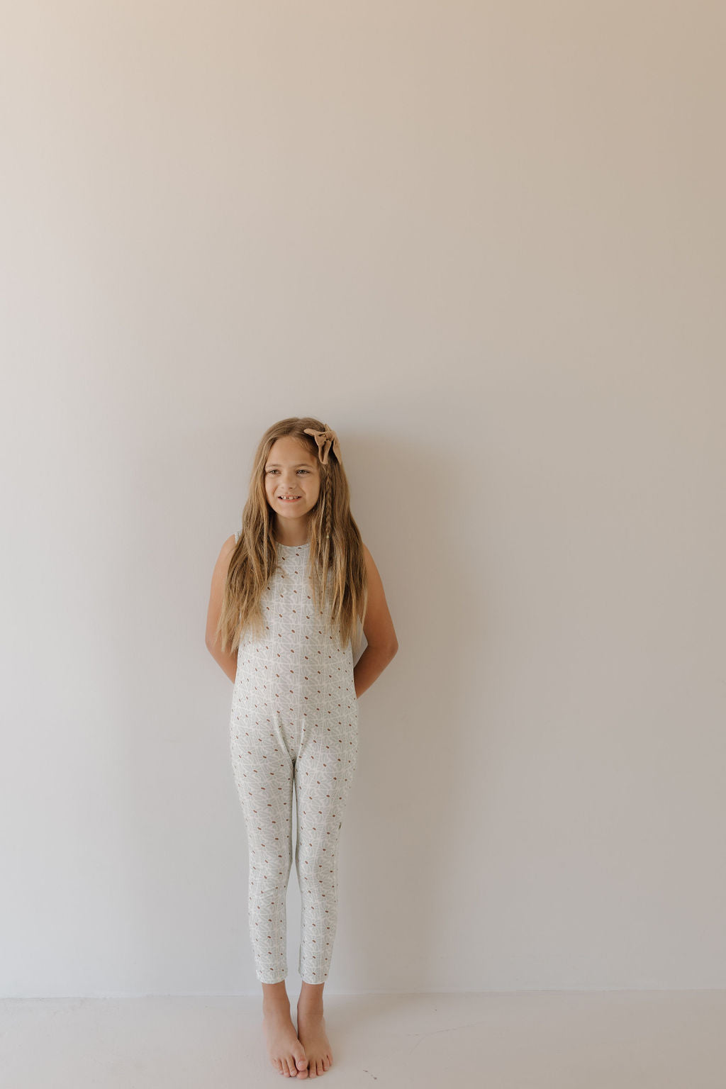 A young girl with long hair stands barefoot against a plain beige wall, smiling gently at the camera. She wears a "Harem Bamboo Romper | Coast" from "forever french baby" and a small bow in her hair.