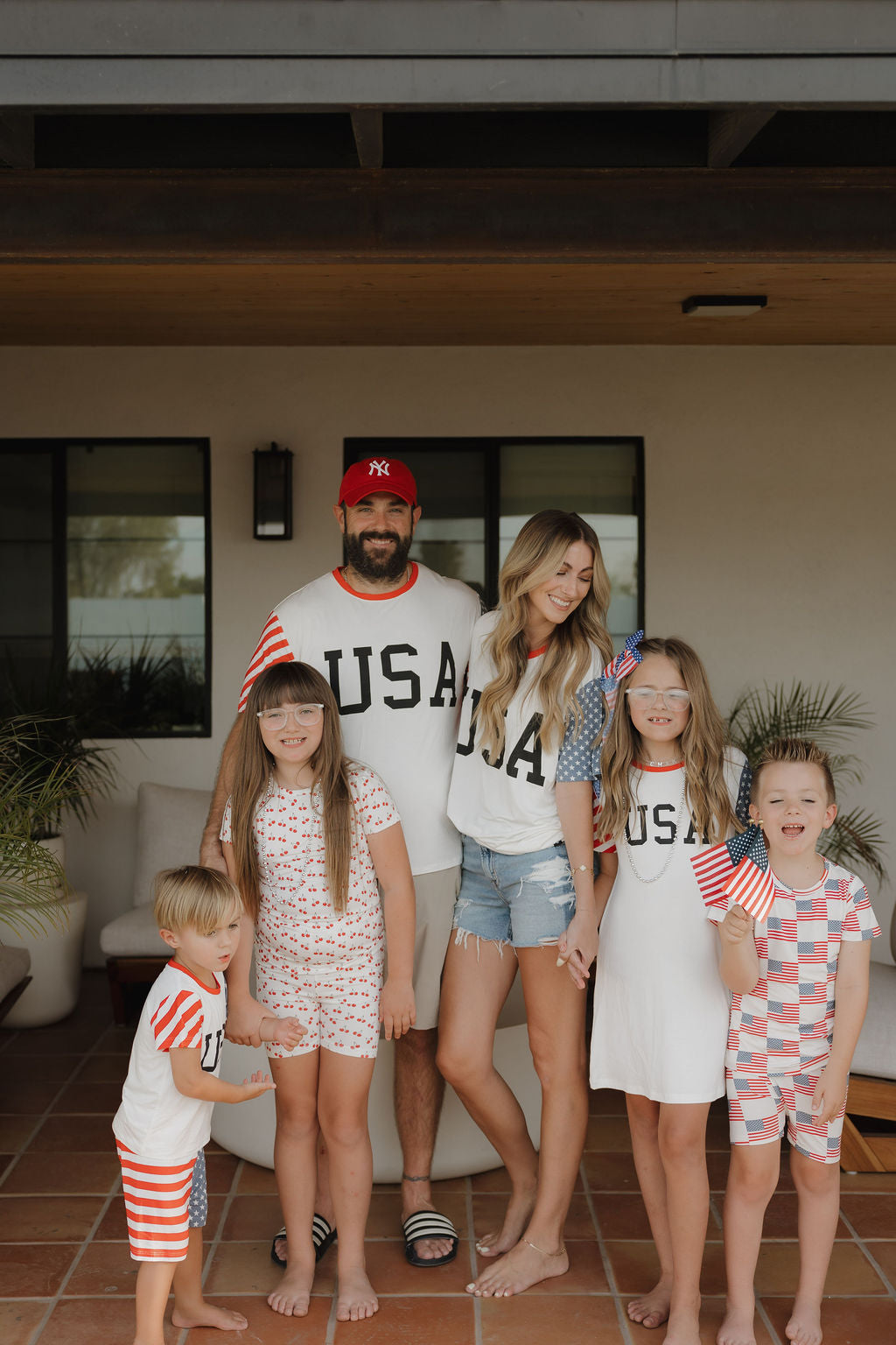 A cheerful family celebrates on the patio, kids holding American flags while wearing forever french baby's Bamboo T-Shirt Dress | Stars & Stripes—a breathable, patriotic choice perfect for festive moments.