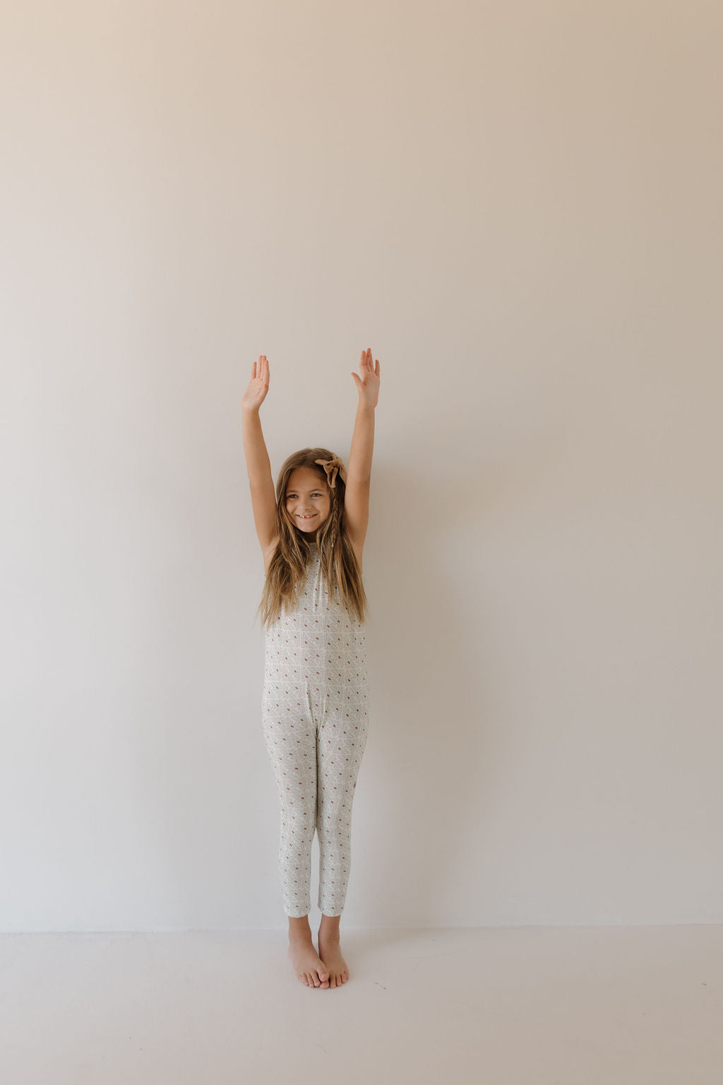 A young girl in a white Harem Bamboo Romper by Forever French Baby stands barefoot against a plain wall, arms raised and smiling.