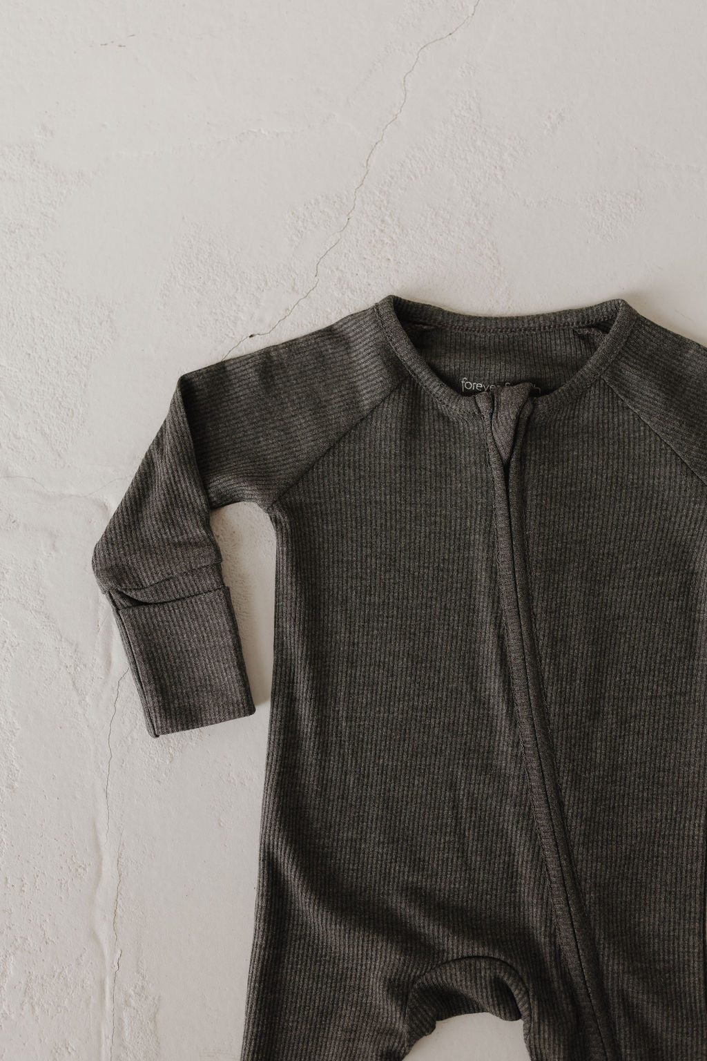 The Bamboo Zip Pajamas | Shadow by forever french baby, a dark gray ribbed onesie made from hypoallergenic bamboo fabric, lies on a textured off-white surface. This breathable garment with a front zipper features a simple yet stylish design.