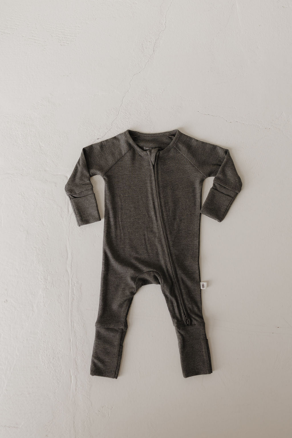 The Bamboo Zip Pajamas in Shadow by forever french baby is a dark gray, long-sleeve onesie laid flat on a light surface. Crafted from hypo-allergenic fabric, it has a visible front zipper and textured design, offering cozy breathability and comfort for infants.